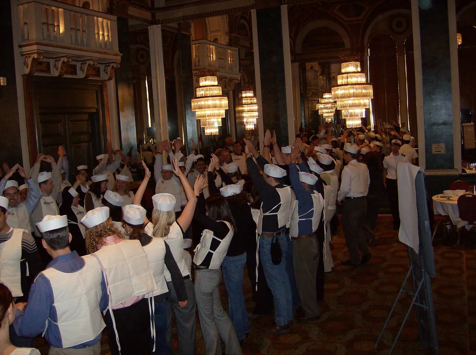 A large corporate team building event in a grand ballroom — hundreds of participants with hands raised during the Save the Titanic experience